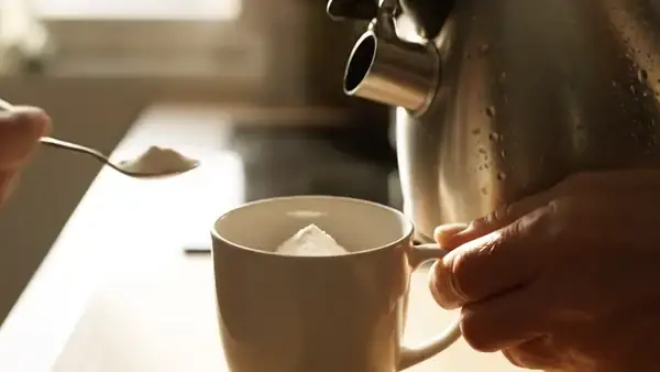 A man's hands pouring hot water from a kettle over pale powder in a mug and stirring it